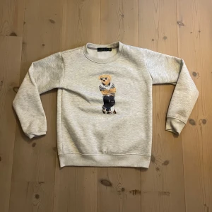 Grå Polo Bear tröja från Ralph Lauren - Superfin ljusgrå sweatshirt från Polo Ralph Lauren med ikoniska Polo Bear-trycket framtill. Tröjan har rund hals, långa ärmar och mjukt material i bomullsmix. Perfekt för dig som gillar klassisk men lekfull stil.