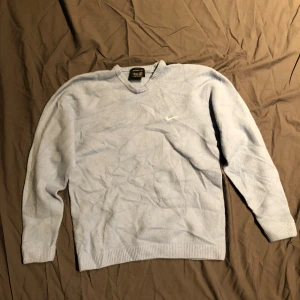 Ljusblå stickad tröja från Nike Golf - Ljusblå stickad tröja från Nike Golf med broderad vit swoosh på bröstet. Klassisk v-ringning och ribbade muddar vid ärmslut och nederkant. Perfekt för dig som gillar sportig och clean stil.