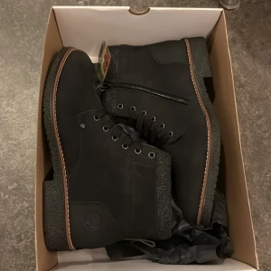 Svarta boots från Rieker med ullfoder - Svarta boots från Rieker med snörning, dragkedja på sidan och mjukt ullfoder. Ovandel i syntet med detaljer i textil och grov sula för extra grepp. Perfekta för kalla dagar med sin varma insida och stilrena look.