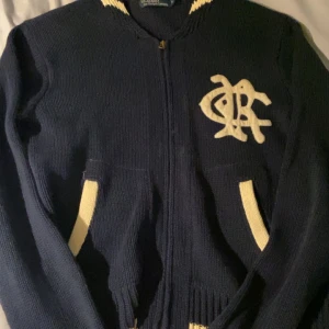 Marinblå kofta från Polo Ralph Lauren - Säljer en marinblå stickad kofta från Polo Ralph Lauren med dragkedja framtill, vita detaljer vid fickor och krage samt ett stort broderat emblem på bröstet. Perfekt för dig som gillar klassisk college-stil och vill ha något som sticker ut. Skick 8/10