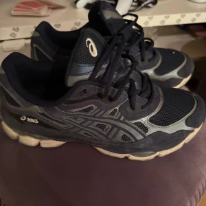 Svarta Asics sneakers med meshdetaljer - Säljer ett par svarta Asics sneakers med grå och vita detaljer. Skorna har meshpaneler för extra andningsförmåga och klassisk snörning. Sulan är vit med svarta inslag och ger bra dämpning. Perfekta för dig som gillar sportig stil.