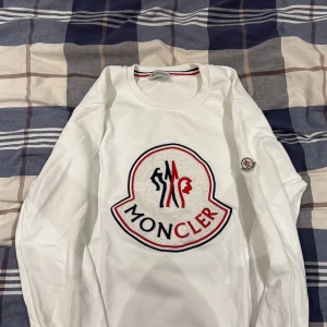 Vit långärmad Moncler tröja - Vit långärmad tröja från Moncler med stort broderat logomärke i rött och marinblått på bröstet. Rund hals och klassisk passform. Moncler-patch på vänster ärm. Perfekt för dig som gillar exklusiva streetwear-vibbar.
