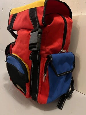 Färgglad ryggsäck från Backsack - Säljer en cool ryggsäck från Backsack i rött, blått, gult och svart. Väskan har flera fickor med dragkedja, justerbara axelremmar och praktiska spännen. Perfekt för dig som gillar färg och vill ha plats för allt du behöver under dagen.