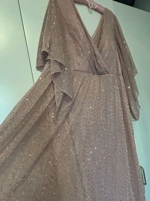 Champagnefärgad glittrig långklänning - Säljer en elegant champagnefärgad långklänning med glittriga detaljer och v-ringad omlottfront. Klänningen har vida, svepande ärmar och ett skirt, glansigt material som ger ett lyxigt intryck. Perfekt för dig som vill sticka ut med en glittrig look.