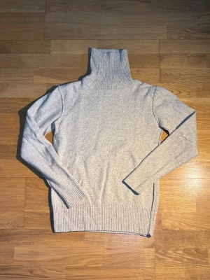 Beige polotröja från East West - Säljer en stilren beige polotröja från East West i storlek M. Tröjan har en hög ribbad polokrage, långa ärmar och breda ribbade muddar vid ärmslut och nederkant. Perfekt för lager-på-lager och chill höst- och vinterdagar.