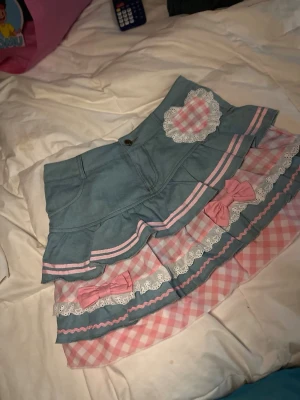 Söt kortkjol med volanger och rosetter - Supersöt kortkjol i ljusblått med rosa och vita detaljer. Kjolen har volanger, rosa rosetter och ett rutigt mönster i rosa och vitt. Dekorerad med spets och ett hjärta framtill.  Det stod att den var storlek S fast den större i stoleken.  den är testad