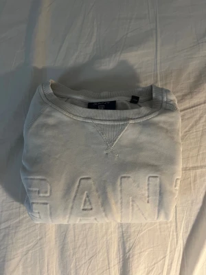 Vit sweatshirt från GANT - Säljer en stilren vit sweatshirt från GANT med broderad logga framtill. Tröjan har rund halsringning, ribbade muddar vid ärmslut och nederkant samt lång ärm. Perfekt för dig som gillar klassisk och enkel stil.