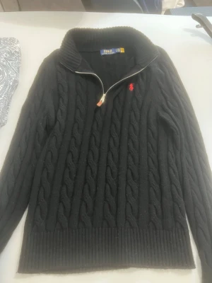 Svart stickad tröja Polo Ralph Lauren - Svart kabelstickad tröja från Polo Ralph Lauren med halv dragkedja och röd broderad logga på bröstet. Tröjan har ribbad krage, muddar och nederkant, samt långärmad passform. Perfekt för dig som gillar klassisk stil med en modern touch.