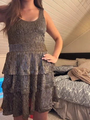 Glittrig guld kortklänning med volanger - Supersnygg glittrig guldklänning med volanger och plisserad struktur. Klänningen är ärmlös och har en rundad halsringning. Den har flera lager med volanger som ger en cool och festlig look. Perfekt för dig som vill sticka ut och glänsa på nästa event!