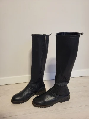 Svarta höga skinnstövlar med dragkedja - Snygga svarta stövlar i skinn med högt skaft och grov sula. Stövlarna har dragkedja på sidan och justerbar snörning upptill för bättre passform. Perfekta till höst och vinter när du vill ha både stil och funktion. Made in Italy.