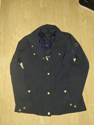 Mörkblå jacka från Peak Performance - Säljer en mörkblå jacka från Peak Performance med guldiga knappar och dragkedja. Jackan har flera fickor framtill och ett Peak Performance-märke på ärmen. Perfekt för dig som vill ha en stilren och funktionell jacka till vardags.