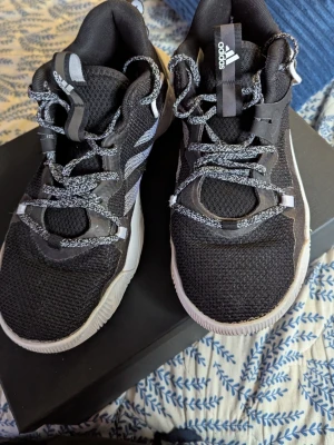 Svarta Adidas sneakers strl 42 - Säljer ett par svarta Adidas sneakers med vit sula och stickad ovandel. Skorna har snörning med svartvita skosnören och coola detaljer på sidan. Märket syns tydligt på plösen och sidan. Perfekt för dig som gillar sportig stil och bekväma skor.