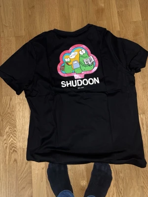 Svart t-shirt från Shudoon - Cool svart t-shirt från Shudoon med färgglad tecknad grafik och text på ryggen, samt liten logga på bröstet. Rund hals och korta ärmar. Materialet känns mjukt och skönt, troligen bomull. Perfekt för dig som gillar unika prints och streetwear-vibe.