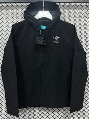 Svart vindjacka från Arc'teryx - Finns i alla storlekar Svart vindjacka från Arc'teryx med huva och snygg logga på bröstet. Jackan har dragkedja framtill och justerbara snören vid huvan. Materialet är lätt och känns sportigt, perfekt för dig som gillar att vara aktiv utomhus.