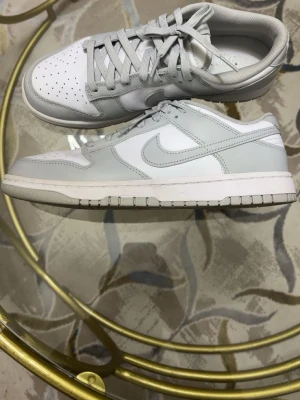 Nike Dunk Low grey - Nike Dunk Low sneakers i fräsch vit och ljusgrå färg. Skorna har klassisk snörning, Ovandelen är i skinn med ljusgrå overlays och vit bas, samt matchande ljusgrå sula. Perfekt för dig som gillar stilrena sneakers med ikonisk design.