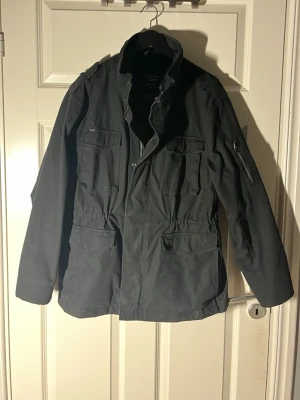 Svart fieldjacket från Superdry - Svart fieldjacket från Superdry med flera fickor framtill och dragkedja på ärmen. Jackan har hög krage, justerbar midja och är tillverkad i slitstark polyester. Perfekt för dig som gillar och vill ha en jacka med coola detaljer. Den är storlek L men passar mig som bär M jackor vanligtvis vid frågor är det bara o skriva// Almen