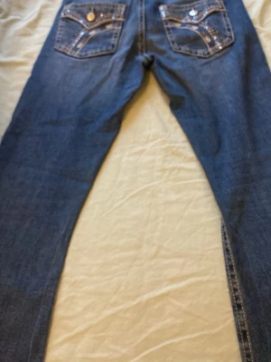 Blå jeans med broderade fickor Gina Tricot - Snygga blå jeans från Gina Tricot i mediumstorlek med unika broderade detaljer på bakfickorna och kontrastsömmar. Jeansen har klassisk femficksdesign och dekorativa knappar på fickorna. Perfekt för dig som vill sticka ut med coola detaljer. Men de är lite sönder vid jyllfen. 170 cm