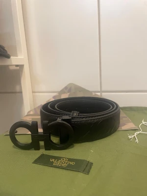 115cm - Bilderna visar ett svart bälte från Salvatore Ferragamo med en stor, rundad metallspänne och mönstrad läderrem. Ingen väst syns på bilderna.