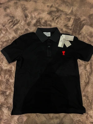 Svart pikétröja från Ami - Svart pikétröja från Ami med klassisk krage och knappar framtill. Tröjan har ett rött broderat hjärta med A på bröstet och Ami-logga broderad i nacken. Materialet är mjuk bomull och modellen är normal passform. Perfekt för en clean och stilren look.