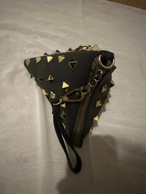 Grå kuvertväska med nitar - Triangelformad grå kuvertväska i skinn med guld- och silverfärgade pyramidnitar över hela ytan. Väskan har en metallkedja, karbinhake och dragkedja samt en handledsrem. Perfekt för dig som gillar edgy detaljer och vill sticka ut.