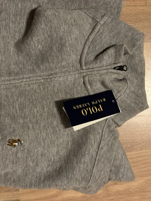 Polo Ralph lauren half zip - Snygg grå långärmad tröja från Polo Ralph Lauren med half zip och ribbade muddar. Tröjan har det klassiska broderade Polo-logot på bröstet och är tillverkad i mjuk bomull. Perfekt för chill dagar och enkel att matcha med jeans eller joggers.