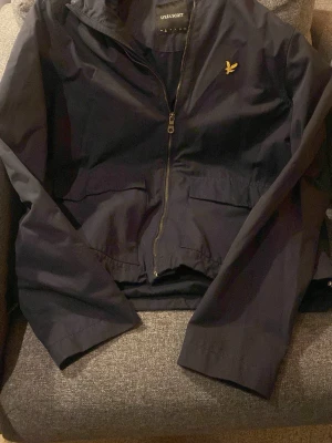 Svart vindjacka från Lyle & Scott - Säljer en mörkblå vindjacka från Lyle & Scott med klassisk gul logga på bröstet. Jackan har dragkedja framtill, två stora fickor och hög krage. Materialet är lätt och smidigt, perfekt för blåsiga dagar. Enkel och stilren design som funkar till det mesta. Ordinarie pris 750. Jackan är helt oanvänd!🤩👌