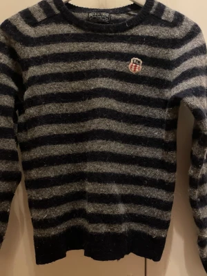 Randig stickad tröja Ralph Lauren - Säljer en stickad tröja från Polo Jeans Company, tillverkad av italienskt garn. Tröjan har mörkblå och grå ränder, rund hals och ett RL-märke på bröstet. Perfekt för dig som gillar klassisk streetstil och vill ha något mjukt och archive