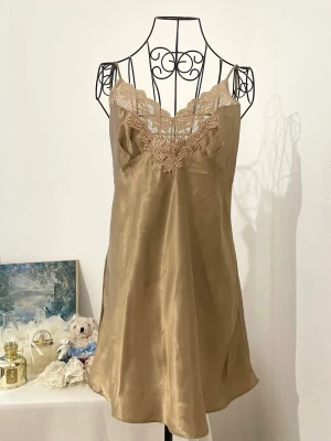 Vintage champagnefärgad slipklänning från Victoria's Secret - Säljer en champagnefärgad nattline med tunna axelband och vacker spetsdetalj vid urringningen. Vintage Gold Tag av Victoria's Secret. Bra skick, dock finns det vissa små märken/punkter på klänningen (se bild 4,5). Storlek: M, passar bäst 38. Dockans storkek: 34