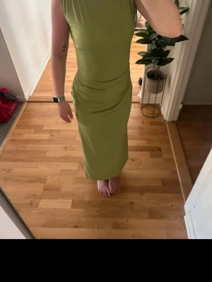 Ljusgrön långklänning från NA-KD - Säljer en snygg ljusgrön långklänning från NA-KD i storlek XS. Klänningen har ett tight passform, draperade detaljer vid midjan och en öppen rygg med slits bak. Materialet är mjukt och stretchigt, perfekt för dig som gillar stilrena och trendiga plagg.