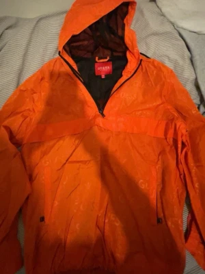 Orange vindjacka från Guess XXL - Snygg orange vindjacka från Guess med halv dragkedja och huva. Jackan har diskret logomönster över hela ytan, meshfoder och praktiska fickor. Perfekt för dig som gillar streetwear och vill sticka ut med färg. Passar dig som vill ha en loose fit.