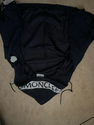 Svart vindjacka från Moncler  - Säljer en svart vindjacka från Moncler . Jackan har huva med stor Moncler-logga, dragsko i huvan och nederkant samt elastiska muddar vid ärmarna. Tillverkad i lätt polyester, perfekt för blåsiga dagar. Klassisk och stilren design med snygga detaljer.