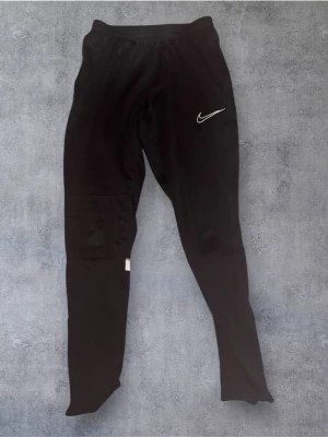 Svarta Nike Dri-FIT träningsbyxor XS - Svarta träningsbyxor från Nike i Dri-FIT-material, med vit broderad swoosh på låret och reflexdetaljer på baksidan av benen. Byxorna har elastisk midja och är tillverkade i 100% polyester. Perfekta för sport eller chill.