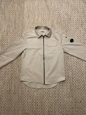 Beige overshirt från C.P. Company - Stilren beige overshirt från C.P. Company med svart dragkedja framtill och klassisk krage. Bröstficka med lock och ikonisk linsdetalj på ena ärmen. Perfekt lager för en clean och modern look.