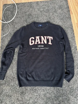 Svart tröja från GANT med tryck - Svart långärmad tröja från GANT med stort broderat tryck på bröstet där det står 'GANT 1949 New Haven, Connecticut'. Klassisk rund halsringning och ribbade muddar. Perfekt för dig som gillar stilrena collegevibbar.