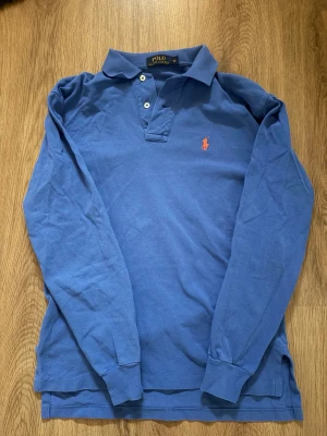 Ralph Lauren Piké - Säljer en klassisk blå långärmad pilé från Polo Ralph Lauren i storlek XS. Toppen har krage, två vita knappar fram och den ikoniska röda logobroderingen på bröstet. Materialet är mjuk bomull och passformen är normal.