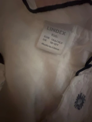 Vit blommig blus från Lindex - Supersöt vit blus från Lindex med mörkblå blommigt mönster och volangdetaljer vid ärmhålen. Klänningen är ärmlös och har en lätt och luftig känsla, perfekt för varma dagar. Passar dig som gillar en lekfull och somrig stil.