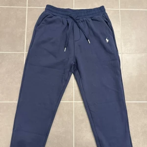 Blå mjukisbyxor från Polo Ralph Lauren - Snygga marinblå mjukisbyxor från Polo Ralph Lauren med klassisk logga broderad på vänster sida. Byxorna har elastisk midja med snörning, sidofickor och en bakficka. Tillverkade i mjukt bomullsmaterial för extra komfort.