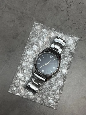 Silver/svart färgad klocka från märket Wzlezanik - Helt ny. Bilderna visar en silvrig armbandsklocka med metallarmband och svart urtavla, men ingen väst finns med. Kan därför inte ge någon västbeskrivning eller matchande taxonomier.