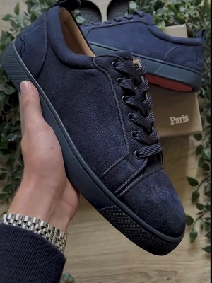 Mörkblå sneakers från Christian Louboutin - Snygga mörkblå sneakers i mocka från Christian Louboutin med klassisk röd sula och broderad logga på hälen. Skorna har rund tå, platt sula och snörning framtill. Perfekt för dig som gillar exklusiva detaljer och stilren design.