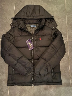 Svart pufferjacka från Polo Ralph Lauren - Svart pufferjacka från Polo Ralph Lauren med huva och röd broderad logga på bröstet. Jackan har två sidofickor med dragkedja och quiltad design. Perfekt för kalla dagar och har en snygg, sportig look.