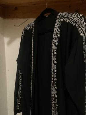 Svart öppen abaya med broderi - Säljer en svart  öppen abaya  med vita broderade detaljer längs kanterna och på ärmarna. Abayan har öppen front och raka, långa ärmar. Perfekt för dig som vill ha något unikt och stilrent i garderoben.