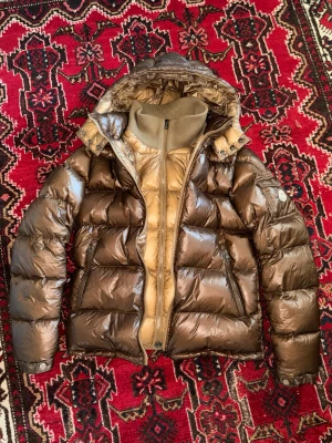 Brun pufferjacka från Moncler - Säljer en brun pufferjacka från Moncler med glansig finish och huva. Jackan har dragkedja framtill, två sidofickor med dragkedja och ribbad krage. Insidan är beige och jackan är fodrad för extra värme. Perfekt för kalla dagar.