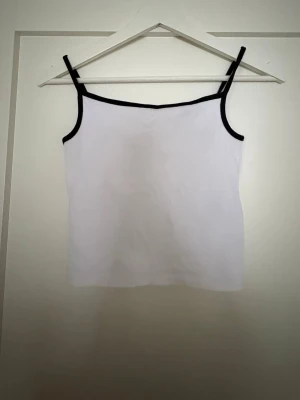 Vit linnetopp med svarta kanter - Säljer en stilren vit linnetopp med svarta kantdetaljer och smala axelband. Toppen har en enkel och rak form, perfekt för varma dagar. Materialet känns mjukt och lätt mot huden. Passar dig som gillar minimalistisk stil och kontraster.
