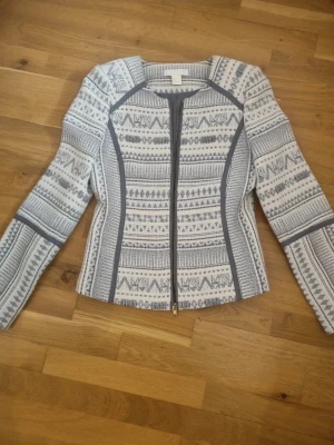 Mönstrad kavajjacka från H&M, strl 36 - Snygg kavajjacka från H&M i storlek 36. Jackan är vit och grå med ett grafiskt mönster och har markerade sömmar i grått. Den har dragkedja framtill och är helfodrad med långärmad passform. Perfekt för dig som gillar stilrena detaljer och mönster.