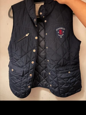 Mörkblå quiltad dunväst Bondelid S - Säljer en mörkblå quiltad dunväst från Bondelid i storlek S. Västen har snygga tryckknappar, en bröstficka med dragkedja och två framfickor med lock. Märkespatch på bröstet och klassisk ståkrage. Perfekt för lager-på-lager och chill stil.