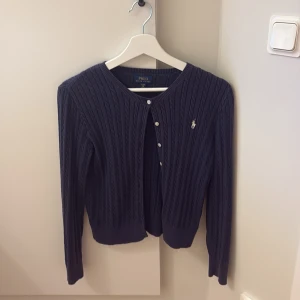 Mörkblå kofta från Polo Ralph Lauren - Det är för 12- 16 år även fast det står XL så är det för barn så den är inte så stor som det värkar. Kabelstickad mörkblå kofta från Polo Ralph Lauren i bomull. Klassisk modell med rund hals, långa ärmar och knappar framtill. Ikoniska loggan broderad på bröstet. Perfekt att slänga över en t-shirt eller klänning.