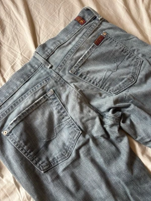 Ljusblå bootcut jeans - Säljer ett par ljusblå bootcut jeans i klassisk denim med fem fickor och snygga sömmar på bakfickorna. Jeansen har en något utsvängd passform nedtill och detaljer i form av små lappar ovanför fickorna. Perfekta för en avslappnad och trendig look.