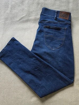 Lee Extreme Motion MVP Slim Taper jeans blå W36 L32 - Mörkblå jeans från Lee i modellen Extreme Motion MVP med Slim Taper-passform. Klassisk femficksmodell i stretchig denim med diskret tvättad effekt, kontrastsömmar och Lee-detaljer framtill och baktill.  Storlek W36 L32 Slim Taper / Slim Fit  Mycket fint begagnat skick med lättare bruksspår och naturlig blekning.  Mått • Midja: ca 46 cm • Grenhöjd fram: ca 28 cm • Ytterbenslängd: ca 107 cm • Innerbenslängd: ca 80 cm • Benvidd vid benslut: ca 19 cm  Material 77% bomull 21% polyester 2% elastan