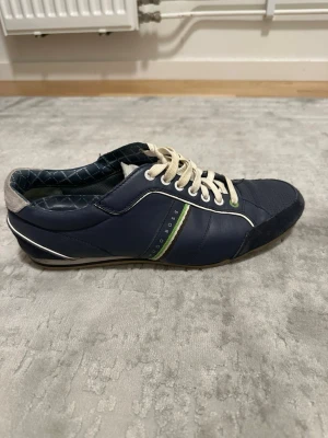 Marinblå sneakers från BOSS - Snygga marinblå sneakers från BOSS med vita snören, gröna och vita detaljer på sidan samt vit häl. Ovandelen är i en mix av läder och textil, och innersulan har ett rutigt mönster. Perfekta för en stilren och sportig look.