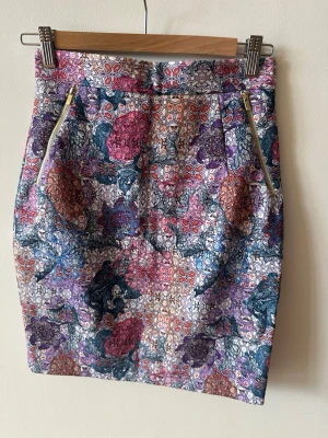 Blommig kjol med dragkedjor - Kort kjol med färgglatt blommigt mönster i lila, rosa, blått och orange. Kjolen har två dekorativa dragkedjor framtill och en figurnära passform. Perfekt för dig som vill sticka ut med en unik och trendig look.